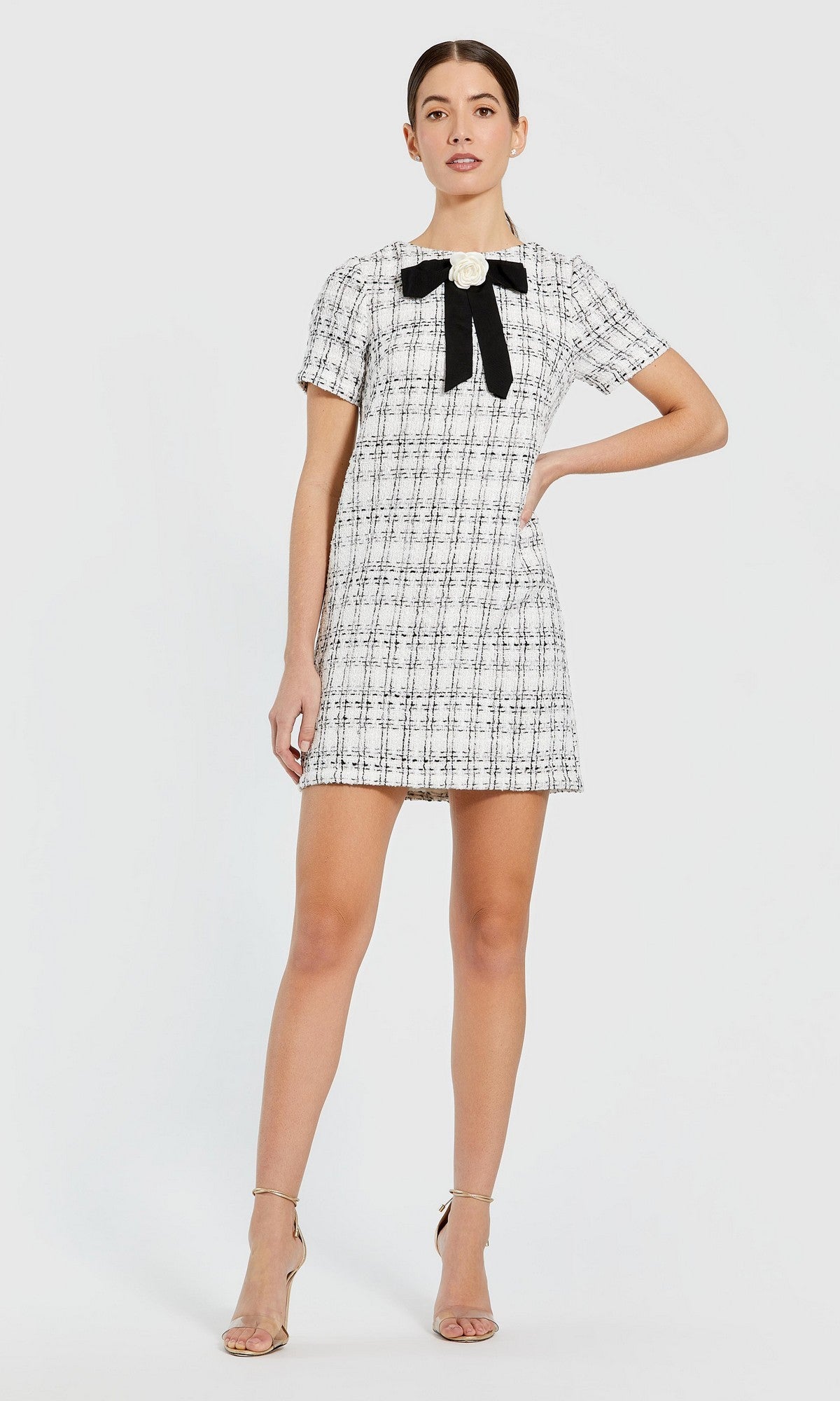 Wearing the Mac Duggal 27796 short semi-formal dress in ivory—a short-sleeve mini silhouette featuring a black-and-white plaid pattern and accented with a large black bow and floral detail at the neckline—a woman stands against a plain background, pairing the look with strappy nude heels.